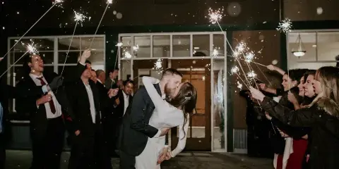 A bride and groom kiss during a sparkler send-off at night while surrounded by guests.