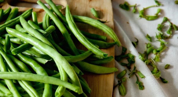 Fresh green beans on a wooden cutting board with trimmed ends on a white cloth.