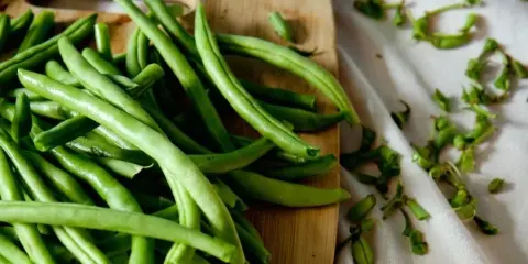 Fresh green beans on a wooden cutting board with trimmed ends on a white cloth.