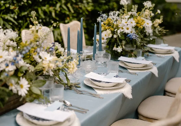 A close-up of a wedding reception table featuring a blue tablecloth, white and yellow floral centerpieces, tall blue candles, and elegant place settings.