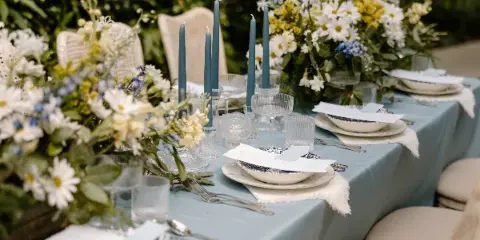 A close-up of a wedding reception table featuring a blue tablecloth, white and yellow floral centerpieces, tall blue candles, and elegant place settings.