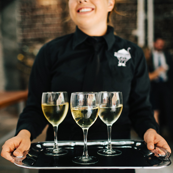 A catering server in a black uniform holding a silver tray with three glasses of white wine.