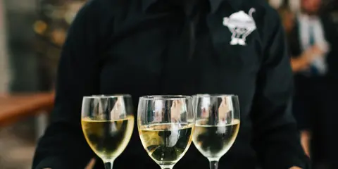 A catering server in a black uniform holding a silver tray with three glasses of white wine.