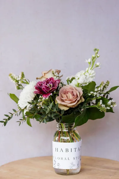A mixed bouquet of white, pink, and mauve flowers with greenery in a glass jar labeled Habitat Floral Studio on a wooden table.