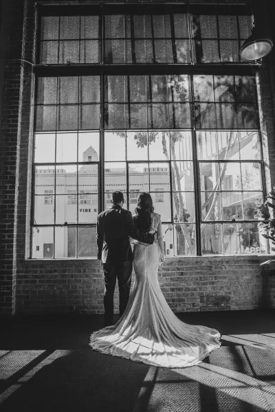 Black and white photograph of a bride and groom standing together in front of a large industrial window.