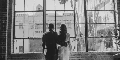 Black and white photograph of a bride and groom standing together in front of a large industrial window.