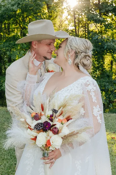 A bride in a white lace gown holding a bouquet and a groom in a tan suit and cowboy hat embracing outdoors.