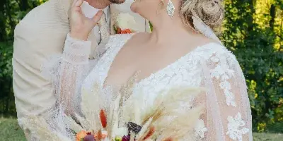 A bride in a white lace gown holding a bouquet and a groom in a tan suit and cowboy hat embracing outdoors.