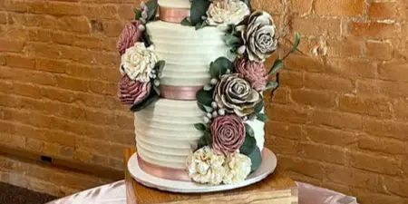 A three-tier white wedding cake decorated with pink and cream flowers, a wooden topper, and a cake knife on a table.