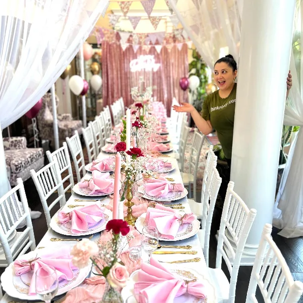 A woman in a green shirt stands beside a long event table set with white chairs, pink napkins, floral arrangements, and balloon decorations.