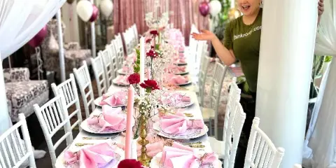 A woman in a green shirt stands beside a long event table set with white chairs, pink napkins, floral arrangements, and balloon decorations.