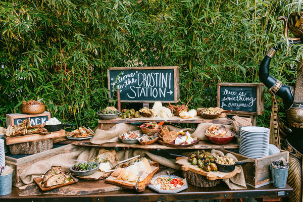 A rustic outdoor catering buffet featuring a crostini station, various prepared dishes, and a snack bar setup.