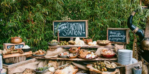 A rustic outdoor catering buffet featuring a crostini station, various prepared dishes, and a snack bar setup.