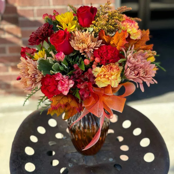 A vibrant autumn-themed floral arrangement featuring red roses, chrysanthemums, and carnations in a glass vase.