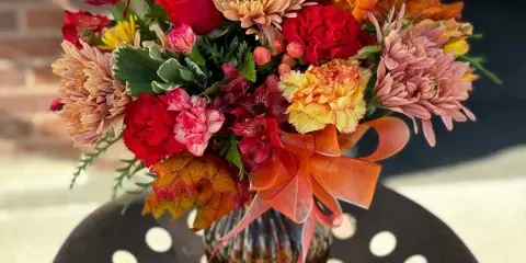 A vibrant autumn-themed floral arrangement featuring red roses, chrysanthemums, and carnations in a glass vase.