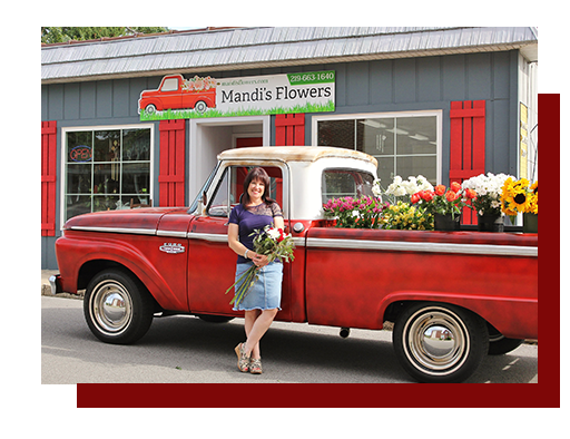 A woman holding a bouquet of flowers stands in front of a red vintage pickup truck loaded with fresh floral arrangements, parked outside the Mandi's Flowers storefront.