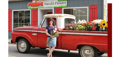A woman holding a bouquet of flowers stands in front of a red vintage pickup truck loaded with fresh floral arrangements, parked outside the Mandi's Flowers storefront.