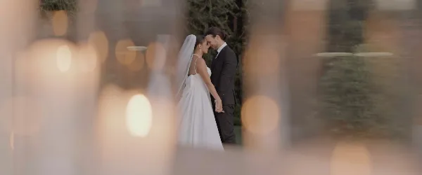 A bride and groom standing close together in an outdoor garden setting, framed by blurred foreground elements.