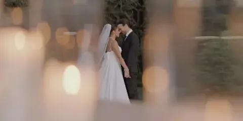 A bride and groom standing close together in an outdoor garden setting, framed by blurred foreground elements.