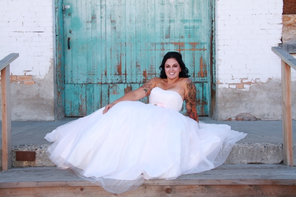 A bride in a white wedding dress sitting on wooden steps in front of a weathered teal door.
