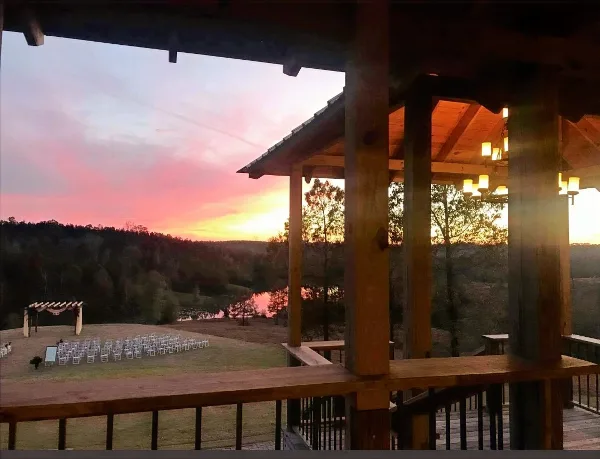 A rustic wooden pavilion overlooking a scenic lake and hills at sunset, with a wedding ceremony setup visible in the foreground.