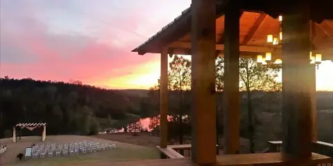 A rustic wooden pavilion overlooking a scenic lake and hills at sunset, with a wedding ceremony setup visible in the foreground.