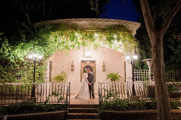 A bride and groom standing in a garden at night in front of a gazebo.