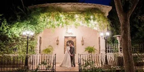 A bride and groom standing in a garden at night in front of a gazebo.