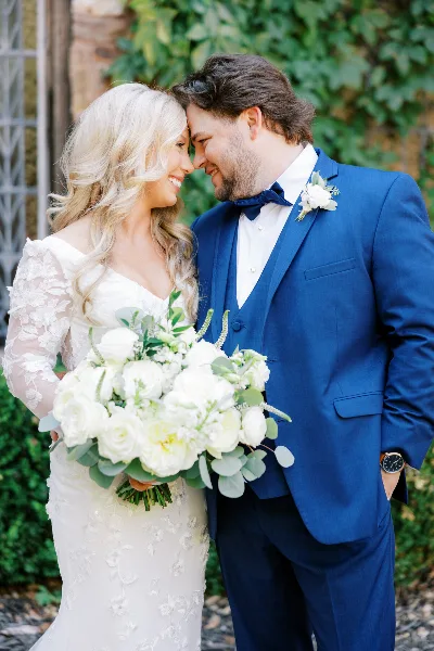 A bride and groom standing together, with the bride holding a large white floral bouquet and the groom wearing a matching white boutonniere.