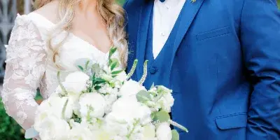 A bride and groom standing together, with the bride holding a large white floral bouquet and the groom wearing a matching white boutonniere.