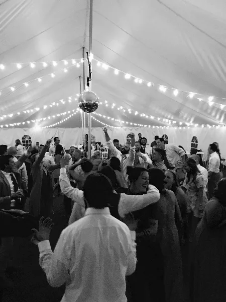Black and white photo of a crowded wedding dance floor with guests dancing under a tent with string lights and a disco ball.