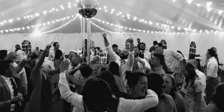 Black and white photo of a crowded wedding dance floor with guests dancing under a tent with string lights and a disco ball.