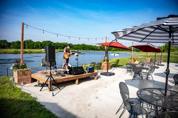 Outdoor event venue with a musician on a stage, string lights, and patio furniture overlooking a lake.