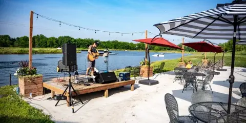 Outdoor event venue with a musician on a stage, string lights, and patio furniture overlooking a lake.