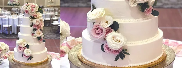 A multi-tiered white wedding cake decorated with pink and white roses and greenery, displayed on a gold stand in a banquet hall.