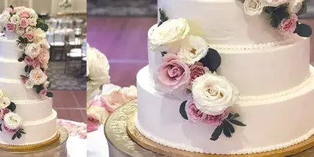 A multi-tiered white wedding cake decorated with pink and white roses and greenery, displayed on a gold stand in a banquet hall.