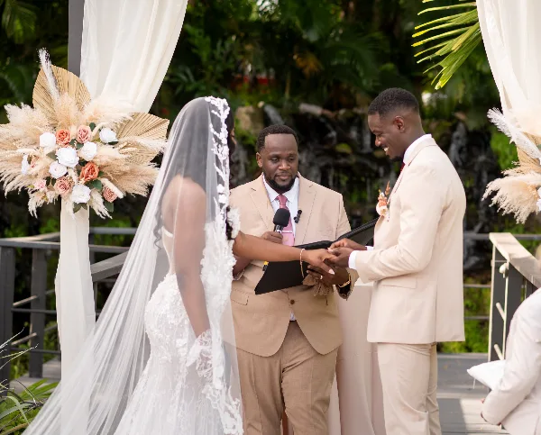 A wedding officiant in a tan suit officiating a ceremony between a bride and groom outdoors.