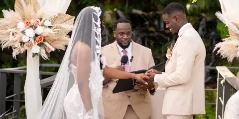 A wedding officiant in a tan suit officiating a ceremony between a bride and groom outdoors.