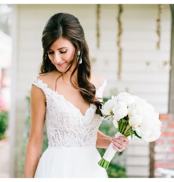 Bride in a white lace wedding dress holding a bouquet of white flowers, with styled hair and makeup.