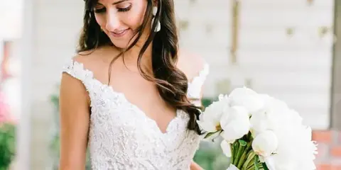 Bride in a white lace wedding dress holding a bouquet of white flowers, with styled hair and makeup.