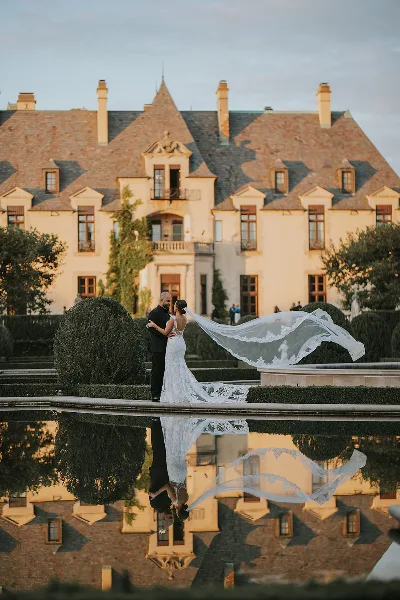 A bride and groom embracing in front of a large European-style estate with a reflecting pool.