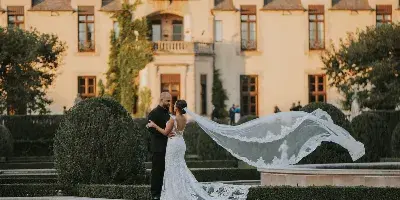 A bride and groom embracing in front of a large European-style estate with a reflecting pool.