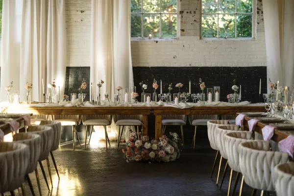 A long wooden dining table set with white linens, floral centerpieces, and candles in a rustic venue with white brick walls.