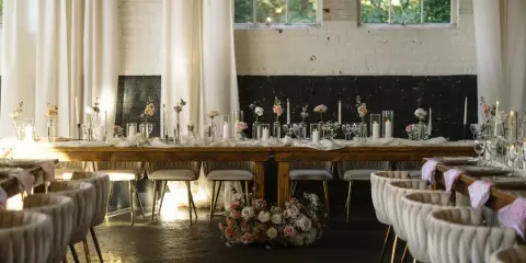 A long wooden dining table set with white linens, floral centerpieces, and candles in a rustic venue with white brick walls.