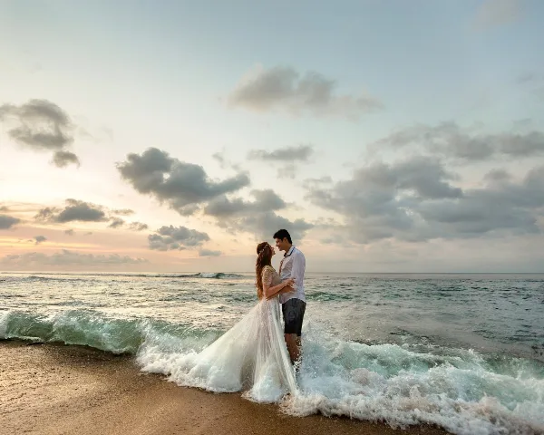 A bride and groom standing on a beach at sunset with ocean waves in the foreground.