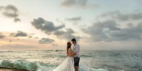 A bride and groom standing on a beach at sunset with ocean waves in the foreground.