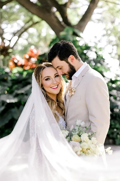 A bride and groom posing together outdoors, with the bride holding a bouquet of white flowers.