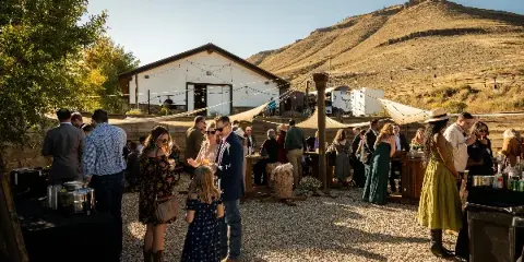 Guests socializing outdoors at a wedding reception in front of a white barn venue with mountain views.