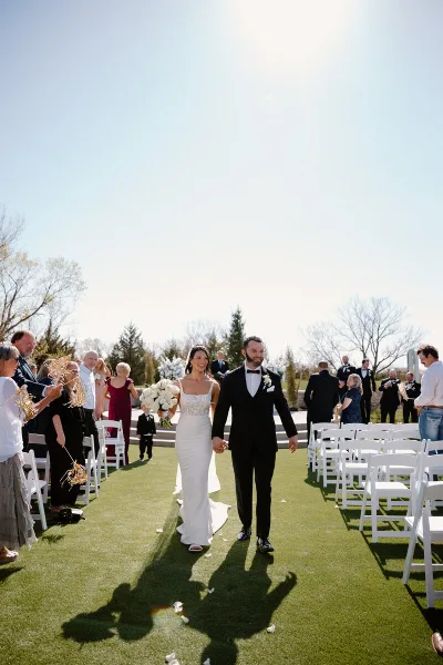 A bride and groom walking down the aisle at an outdoor wedding ceremony, smiling and holding hands.