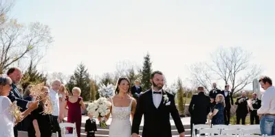 A bride and groom walking down the aisle at an outdoor wedding ceremony, smiling and holding hands.